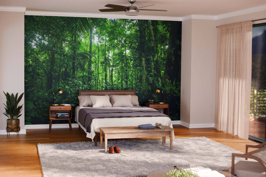 A modern open-plan living and dining room with wooden floors, teal armchairs, and a dark dining table features the Tropical Rainforest Landscape Wall Mural as a lush focal point. Pendant lights hang above the table; large windows bring in natural light.