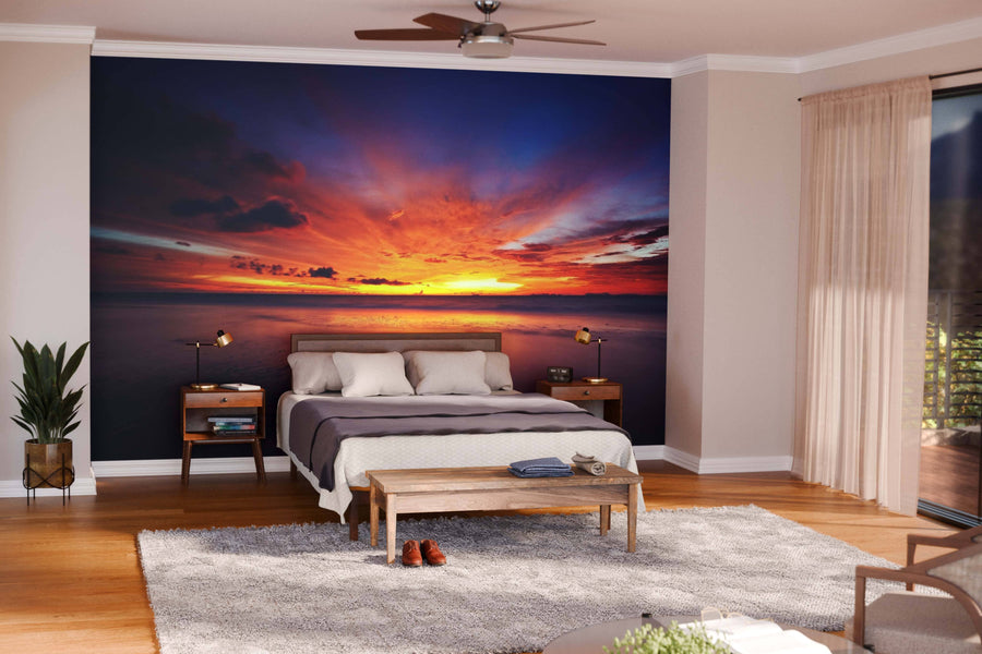 A modern living and dining room with wooden floors and mid-century furniture features the Tropical Sea Sunset Wall Mural, showcasing vivid orange, red, and purple clouds over the ocean as a striking focal point in the open space.