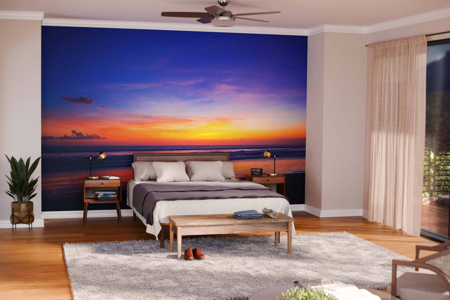 A modern living-dining room with wooden floors, teal chairs, a rug, and a sculpted coffee table features a vivid “Tropical Sunset on the Beach Wall Mural” in orange, purple, and blue tones above calm water on the back wall.