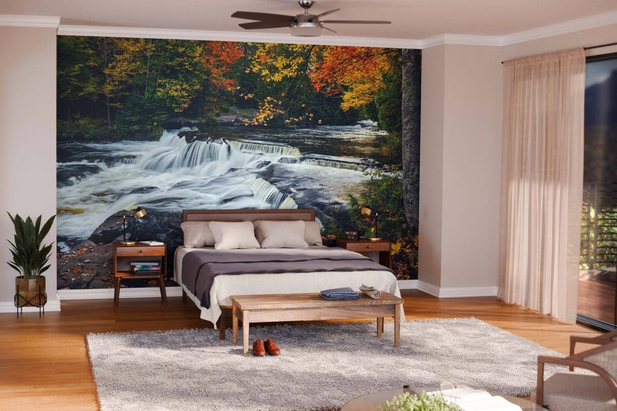 A modern living-dining area features wood floors, teal armchairs, and a dining set. The Upper Bond Falls Wall Mural depicting an autumn forest waterfall adds vibrance, while pendant lights hang above the table.