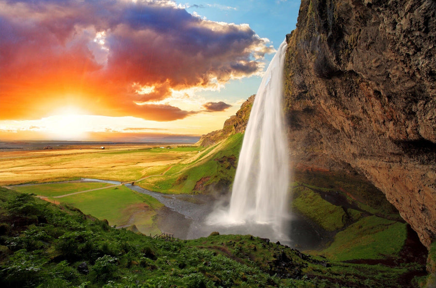 A modern bedroom with the "Waterfall in Iceland Wall Mural" showcases a vivid landscape of cascading water, lush fields, and a sunset. A wooden bed, side tables, greenery, and sheer curtains complete the bright, inviting space.