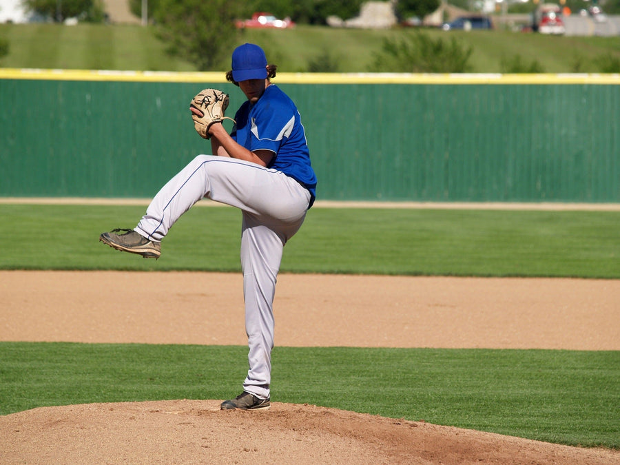 A bedroom with a desk and chair, single bed, soccer ball on the floor, white cabinets, and windows with blinds. The wall features the Winding Up Wall Mural showing a baseball pitcher in a blue uniform.