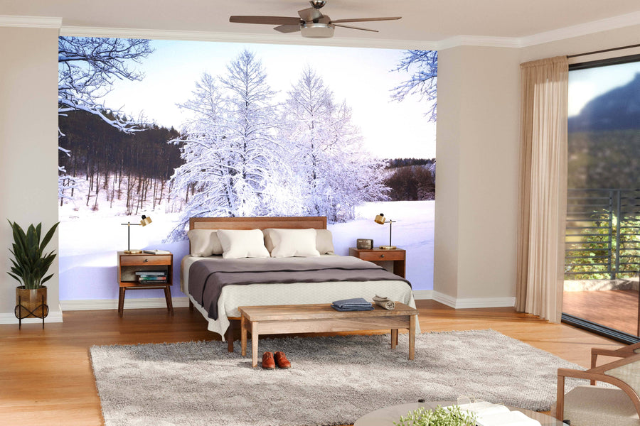 A modern open-plan living and dining room with wooden floors features teal chairs, a gray sofa, and round pendant lights. The focal point is the "Winter Landscape with Snow-Covered Trees Wall Mural," adding cozy, wintry charm.