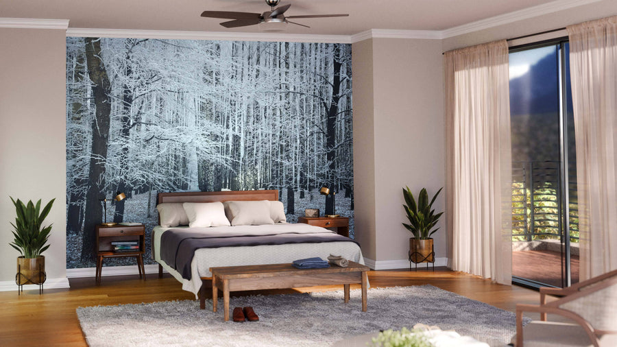 A modern bedroom features a large bed, wooden nightstands, potted plants, and a bench. One wall showcases the Winter Trees Dusting with Snow Wall Mural, while glass doors with sheer curtains lead to a balcony abundant with greenery.