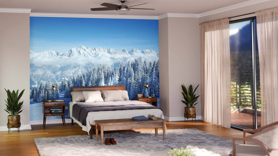 A cozy, modern living room with green and brown furniture features the "Winter in the Alps Wall Mural." Large windows show snowy pines and mountains under a blue sky, while warm lighting contrasts with the wintry scene outside.