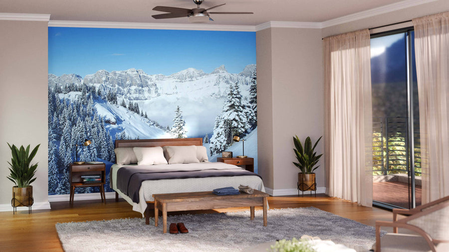 A modern living and dining area with wood floors features blue chairs, a wooden bench, a dining table under pendant lights, and the “Winter in the Swiss Alps Wall Mural,” showcasing snowy mountains beneath a clear blue sky.