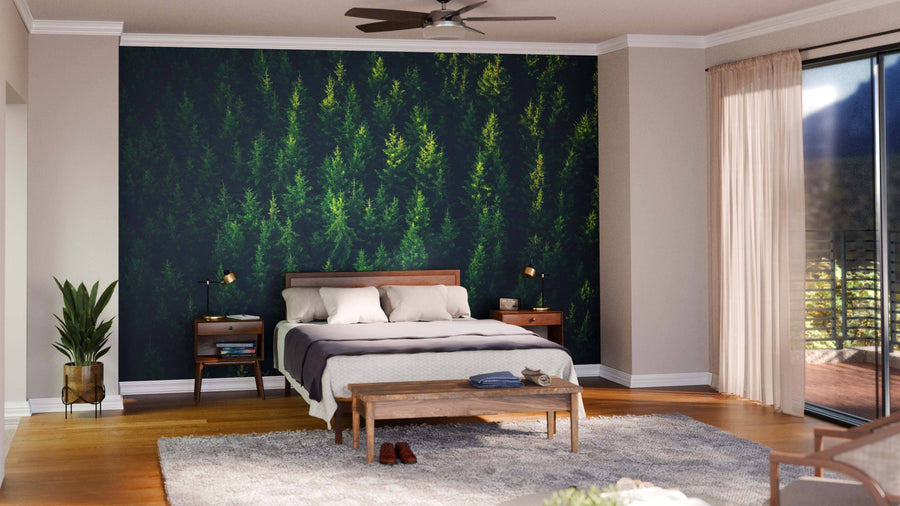 A bedroom features the Above of the Forest Wall Mural behind a wooden bed with white bedding and a gray blanket. Nightstands with lamps flank the bed. A potted plant, bench, rug, and large window with sheer curtains complete the space. Ceiling fan above.