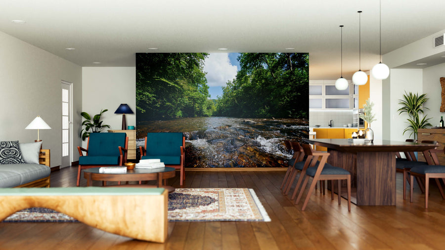 Spacious living-dining room with wood floors, modern furniture, and a large Abrams Creek Wall Mural featuring a river and green trees. Blue chairs, wooden sofa, pendant lights, and plants complete the stylish décor.