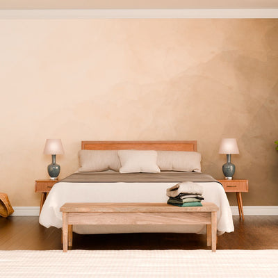 A minimalist bedroom reveals a wooden bed adorned with beige and gray bedding. Two bedside tables hold matching gray lamps. A bench with folded clothes rests at the foot of the bed, while a potted plant stands nearby. Soft light cascades through sheer curtains onto the "A Way Out" wall mural on the right wall.