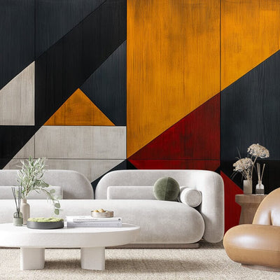 The modern living room is highlighted by the Abstract Contrast Wall Mural, displaying geometric shapes in black, orange, yellow, and red. It pairs well with light gray sofas, a low white oval coffee table with greenery and books, a round brown chair, and a wooden side table adorned with dried flowers.