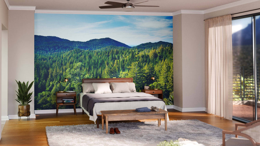 A bright bedroom with wood flooring, beige walls, and large windows features white bedding and gray pillows. The Aerial View Forest Wall Mural adds a scenic, nature-inspired backdrop behind the bed.