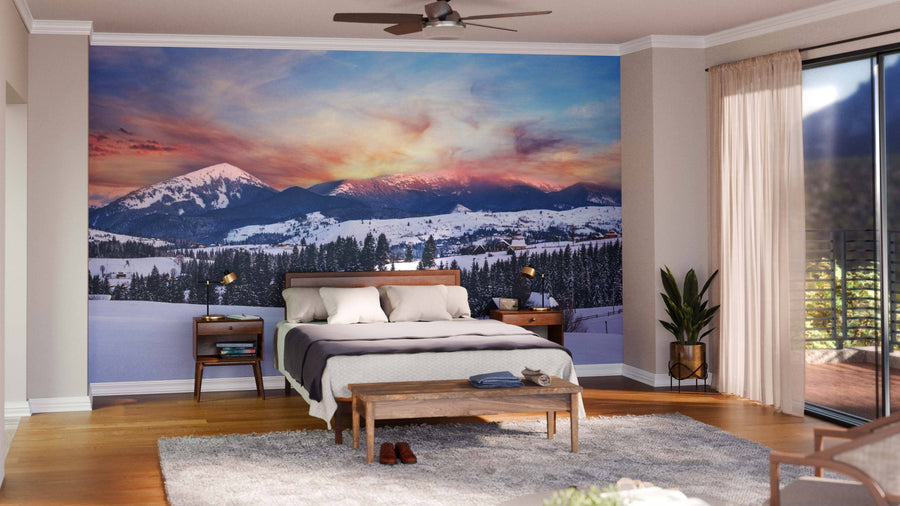 A cozy bedroom features the Alpine Village Outskirts Wall Mural of snowy mountains at sunset behind a bed with white linens, nightstands with lamps, a bench, plant, rug, and sliding glass doors with sheer curtains.