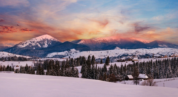 The Alpine Village Outskirts Wall Mural captures a snowy mountain landscape at sunset, with snow-covered peaks, rolling hills, pine trees, and scattered houses beneath a vibrant orange, pink, and purple sky—perfect for showcasing the tranquil beauty of a winter evening in custom printed murals.