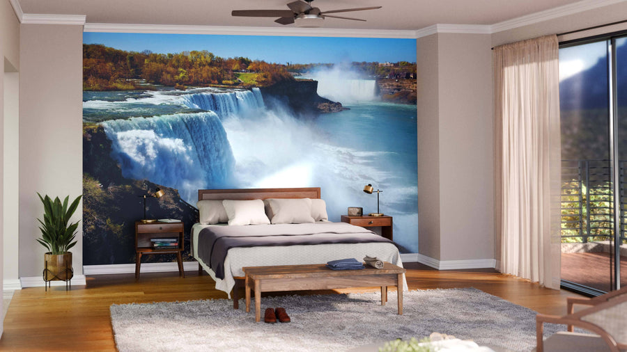 A bright bedroom features the American Side of Niagara Falls Wall Mural behind a wooden bed with white bedding. Nightstands with lamps, plants, a gray rug, a wooden bench, and large windows with sheer curtains complete the space.