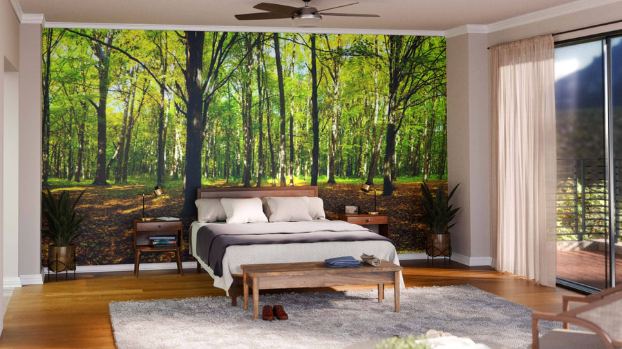 A bedroom with a bed, nightstands, and a bench faces an Autumn Forest Panorama Wall Mural. Wood floors, a gray rug, sliding glass doors with sheer curtains, and natural light create a cozy, nature-inspired space.