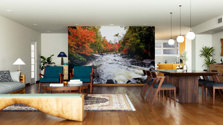 A modern open-concept living and dining room with wooden floors, blue armchairs, plants, contemporary furniture, a wooden dining table and chairs, pendant lights above, and an Autumn Leaves Wall Mural depicting a vibrant river scene.