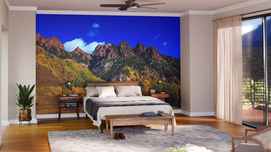 A bright bedroom with wooden floors includes a bed with neutral bedding facing the Autumn Mountain Landscape Wall Mural. At the foot of the bed is a bench, with a plant, rug, and glass doors on the right letting in sunlight.