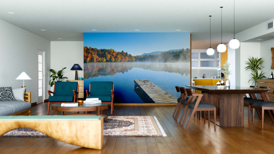 A modern open-plan living and dining area features wood floors, mid-century furniture, and the Autumn Serenity Wall Mural depicting a tranquil lake with autumn trees. Pendant lights hang over a dark wood table; plants and rugs add warmth.