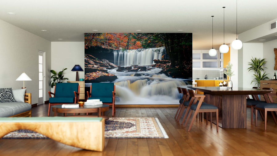 A modern open living-dining room features wood floors, blue chairs, a large Autumn Waterfalls in Park Wall Mural, a long wooden dining table with chairs, pendant lights, a patterned rug, and a cozy, inviting atmosphere.