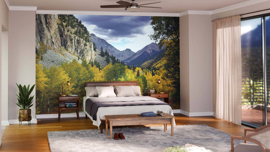 A modern bedroom features a large bed, wooden furniture, houseplants, and a gray rug. Floor-to-ceiling windows with sheer curtains open to a balcony. The far wall showcases the "Autumn in Colorado Wall Mural," displaying a mountain landscape.