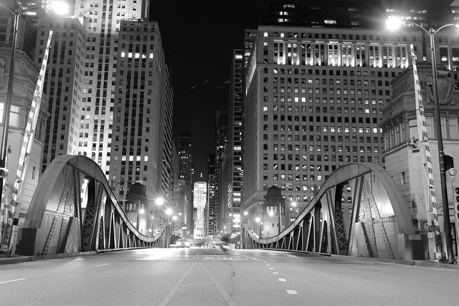 The Chicago Downtown Bridge at Night Wall Mural showcases a well-lit urban scene with an arching bridge leading to a cityscape. The mural features tall buildings, curved beams, overhead lights, red and white barriers, and empty streets—perfect for a stunning custom-printed mural.