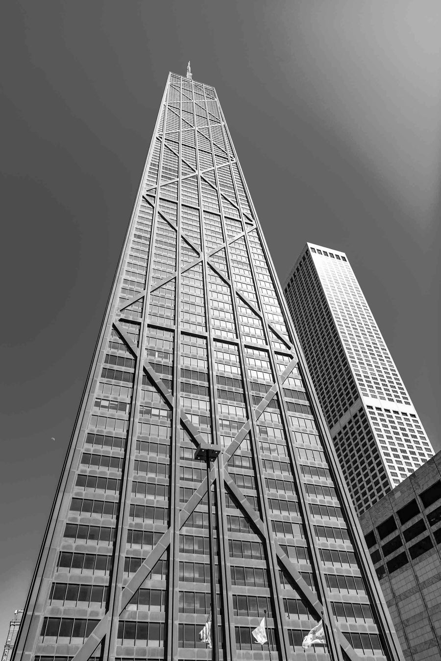 A low-angle view of a skyscraper with a crisscrossed steel framework and reflective glass windows against a clear blue sky. A neighboring building is visible, offering inspiration for the Chicago John Hancock Building Wall Mural, capturing height and modern architectural style.