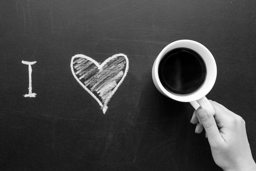 A hand holds a white cup of black coffee against a dark green chalkboard with "I" and a heart in white chalk, forming "I love coffee." The heart resembles art on the Coffee is Love Wall Mural, creating an expressive backdrop.