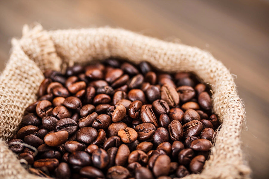 A modern café interior with wooden tables, stools, pendant lights, and sunlit windows features the Bag of Coffee Beans Wall Mural on the back wall. Small plants and tableware decorate the tables for a fresh touch.