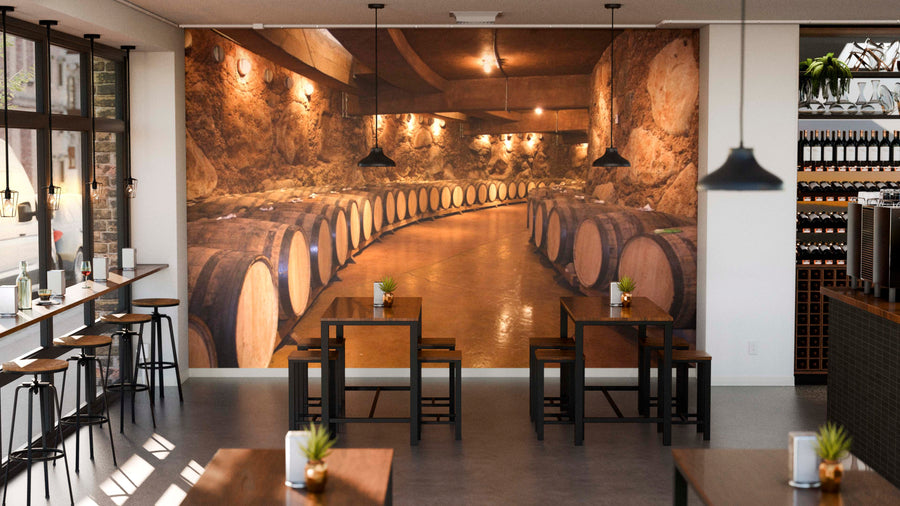 Modern café with wooden tables and stools, indoor plants, and a large Aged Oak Barrels Wall Mural. Pendant lights hang from the ceiling, while shelves lined with wine bottles decorate the right side of the space.