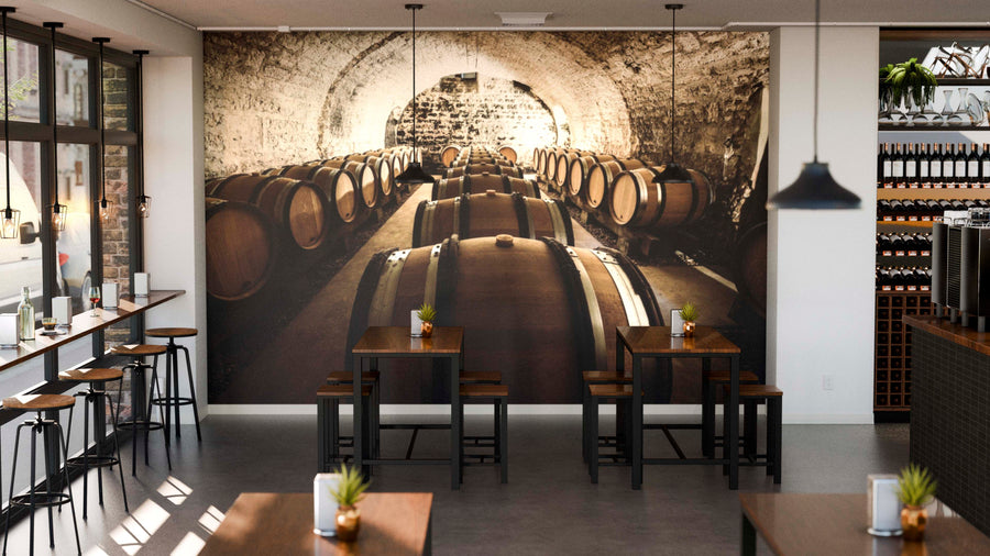 A modern café with dark wood tables and stools features the Aging Cellar Wall Mural—a striking wine cellar scene with wooden barrels—on its back wall. Sunlit windows, potted plants, and shelves of wine bottles complete the charming space.