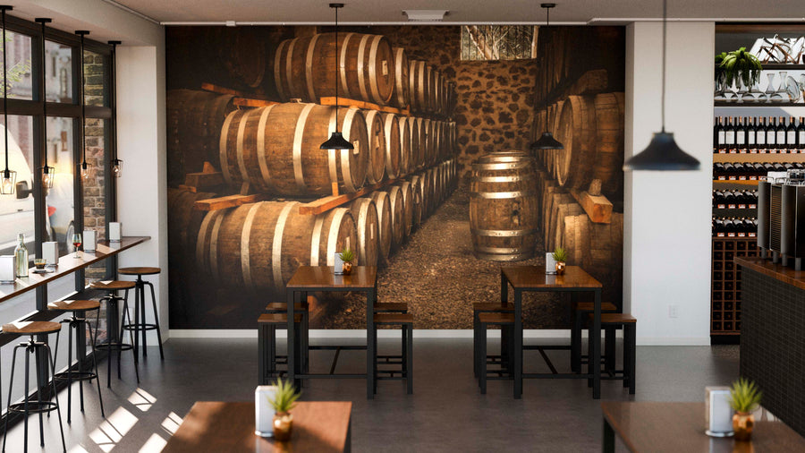 A modern cafe features wooden tables, chairs, potted plants, and hanging lights. One wall showcases the Aging Oak Barrels Wall Mural, depicting stacked barrels in a rustic cellar for a cozy feel. Large windows line the left side.