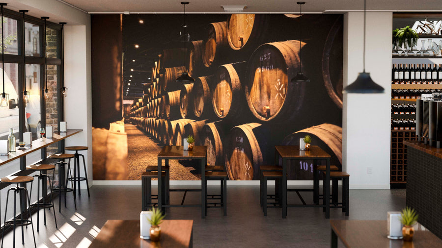 Modern café interior with wooden tables, black chairs, and large windows. The back wall displays the Barrel Storage Wall Mural showing stacked wine barrels. Shelves lined with wine bottles run along the right wall beneath pendant lights.