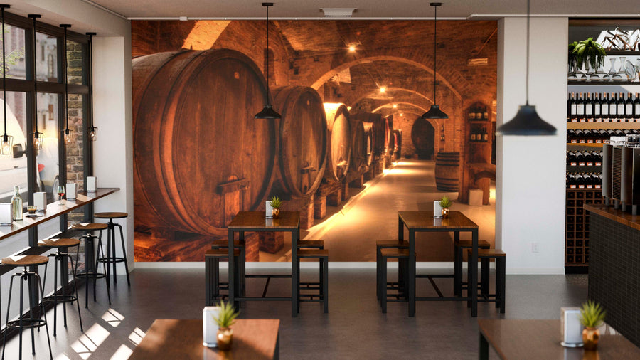 Modern café interior with wooden tables, stools, and plants on each table. The back wall features the Barrel Vault Wall Mural depicting a dimly lit wine cellar with large wooden barrels and arched brick ceilings.