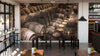 A stylish café interior features wooden tables and chairs, potted plants, and a Barrel Warehouse Wall Mural of stacked barrels on the back wall. Large windows bring in natural light, complemented by pendant lights from the ceiling.