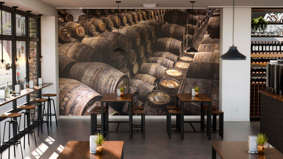 A stylish café interior features wooden tables and chairs, potted plants, and a Barrel Warehouse Wall Mural of stacked barrels on the back wall. Large windows bring in natural light, complemented by pendant lights from the ceiling.
