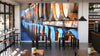 Modern cafe interior featuring the Barrels Wall Mural on the back wall. Black tables and stools are neatly arranged, each adorned with small plants. Wine bottles line the shelves, enhancing the space’s cozy ambiance.