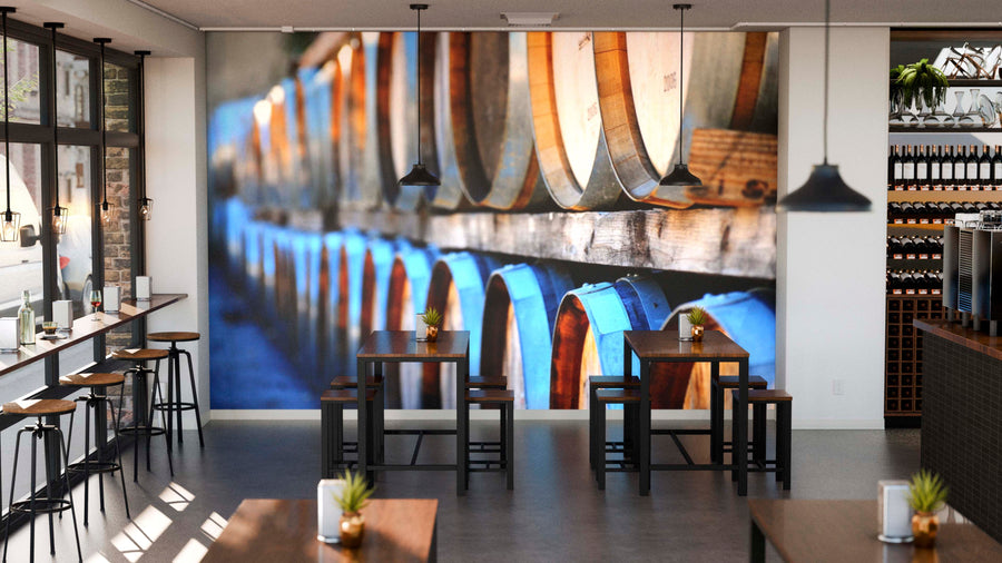 Modern cafe interior featuring the Barrels Wall Mural on the back wall. Black tables and stools are neatly arranged, each adorned with small plants. Wine bottles line the shelves, enhancing the space’s cozy ambiance.