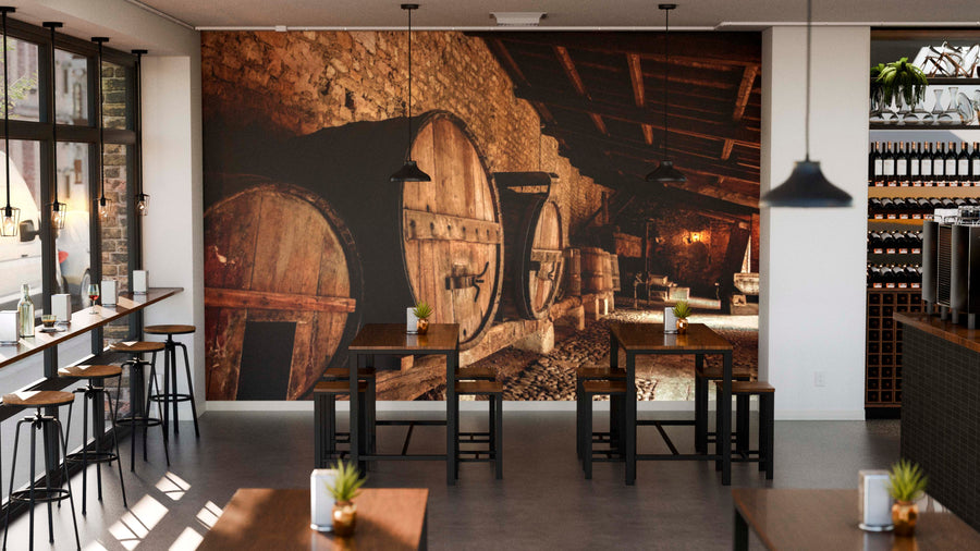 A modern café with wooden tables and black chairs features the "Barrels Lined Against a Stone Wall Wall Mural." Pendant lights hang overhead, potted plants decorate the tables, and shelves with wine bottles line the right side.