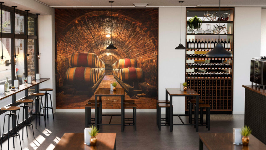 Modern café with wooden tables, stools, wine bottles on shelves, pendant lights, and a large Brick-Walled Wine Cellar Wall Mural featuring barrels. Sunlight streams in as small plants decorate the tables.