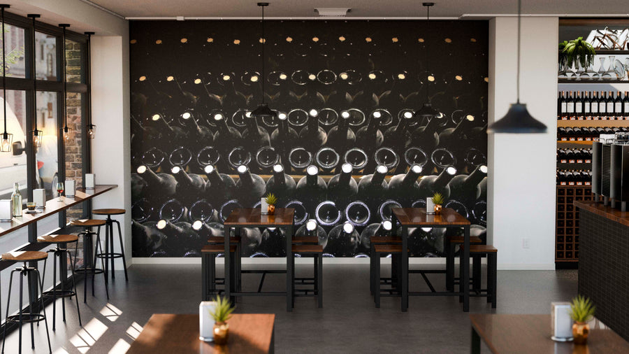 Modern cafe interior features wooden tables, black chairs, small potted plants, and the Cellar Stacked Bottles Wall Mural showing rows of wine bottles. Pendant lights hang above, with shelves and a coffee machine on the right.