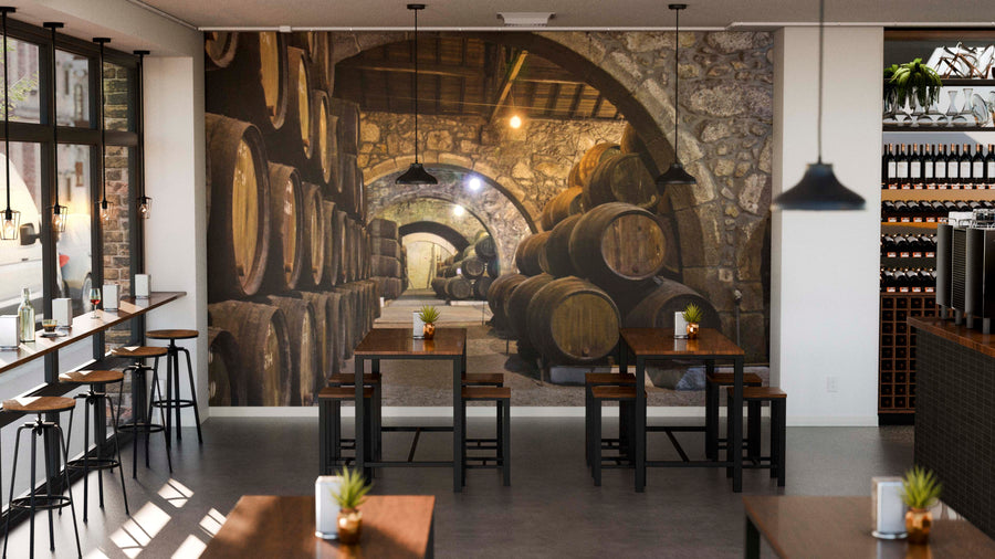 A cozy café with wooden tables and stools faces a large Cellar with Wine Barrels Wall Mural. Pendant lights hang from above, small potted plants sit on tables, and large windows brighten the space with natural light.