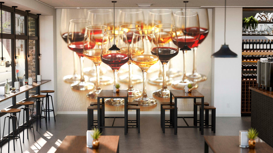 A modern wine bar with tall tables and stools features the Collection of Wine Glasses Wall Mural on its back wall. Sunlight pours through large windows, while wine bottles and small plants accent each table.