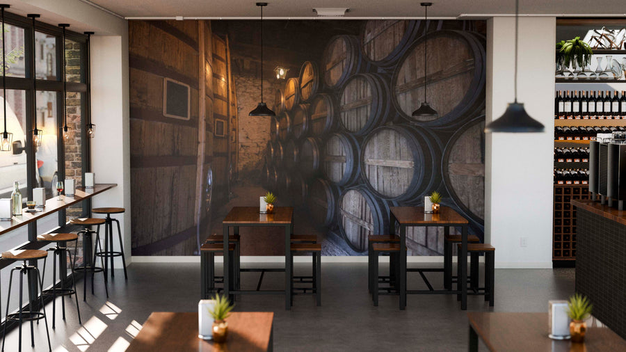 Modern café with wooden tables and stools. A Dimly Lit Cellar Wall Mural featuring stacked wine barrels dominates the back wall. Potted plants and condiments adorn tables, pendant lights hang above, and large windows fill the space with natural light.