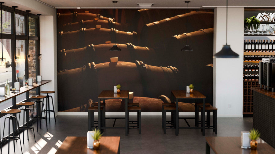 A modern cafe interior boasts wooden tables and chairs, small potted plants, pendant lighting, and a large Dimly Lit Wooden Wine Barrels Wall Mural. Sunlight through windows adds to the cozy, industrial vibe.