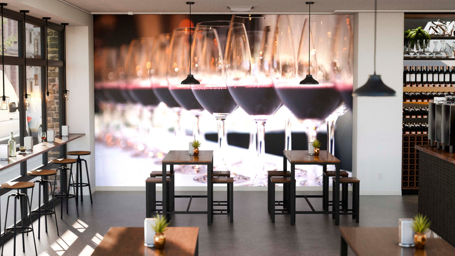 Modern wine bar interior featuring wooden tables, stools, potted plants, pendant lighting, and shelves of wine bottles. The right wall showcases the Glasses of Red Wine Wall Mural. Large windows fill the space with natural light.