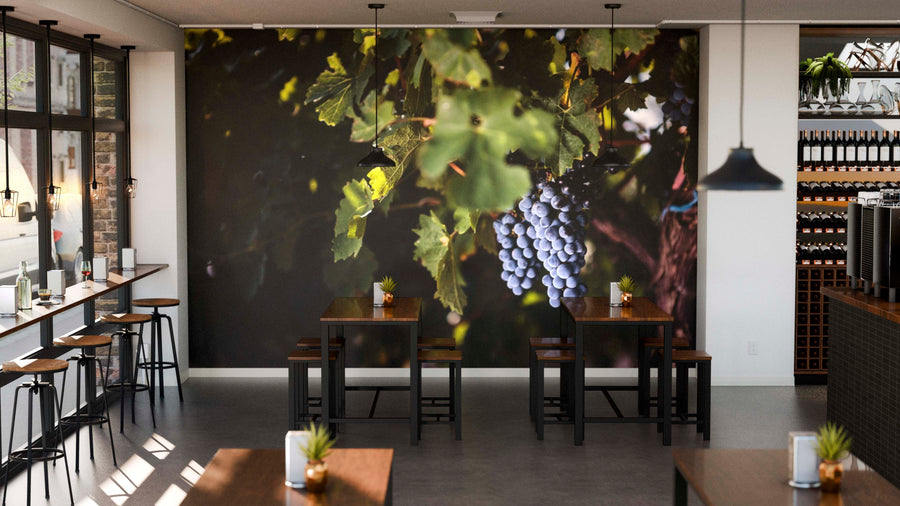 A modern café features wooden tables, black chairs, and potted plants. The "Grapes on a Vine Wall Mural" displays vivid purple grapes. Pendant lights hang above, and wine bottles are neatly arranged on shelves to the right.