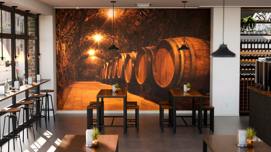 A modern café with wooden tables and stools, decorated with potted plants. The Rustic Wine Cellar Wall Mural depicts a warmly lit underground cellar. Natural light streams in through large windows on the left.