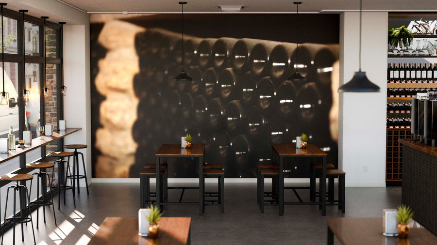 A modern café features the Stacked Wine Bottles Wall Mural on its back wall, with wooden tables and chairs, bar seating by tall windows on the left, pendant lights above, and small potted plants as table centerpieces.