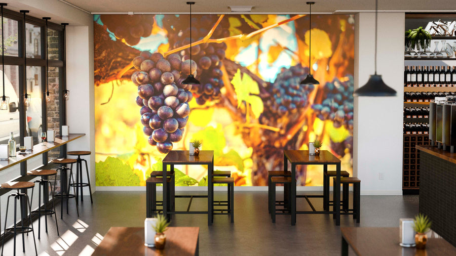 Modern café interior with wooden tables and black chairs, featuring the Sunlit Vineyard Wall Mural—depicting purple grapes and yellow-green leaves—on the back wall. Pendant lights and potted plants complete the décor.