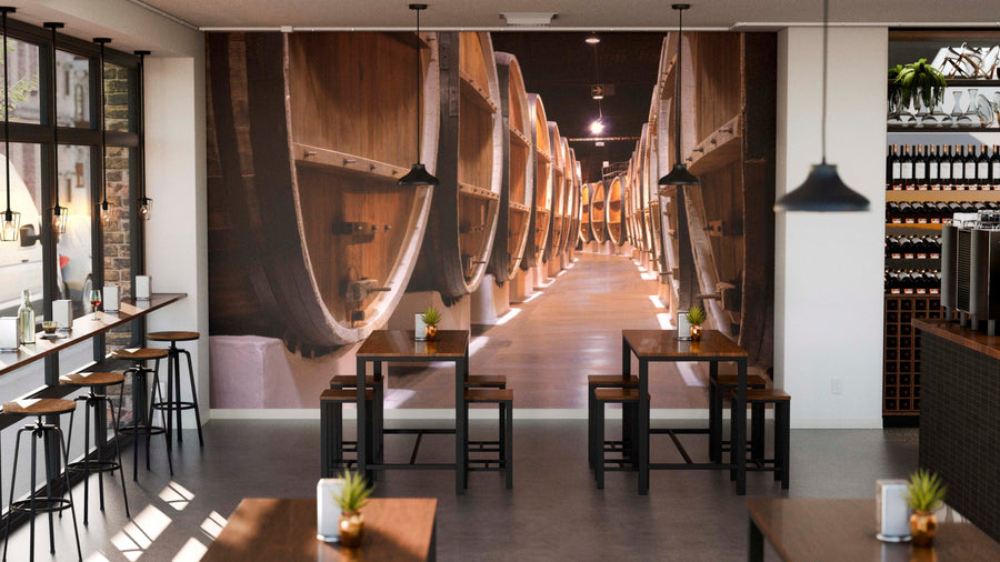 A modern café features wooden tables and stools, potted plants, pendant ceiling lights, a wine rack on the right, and a Timeless Oak Wall Mural showing a row of wine barrels in a cellar as a peel-and-stick accent.